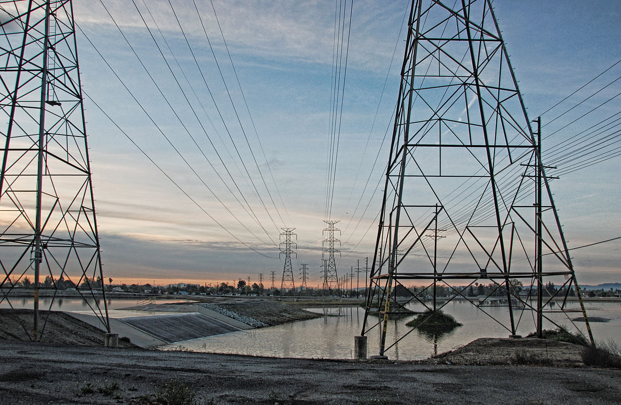 A two-ink risograph print of tall metal transmission towers crossing a calm water channel at dusk, rendered in soft amber and slate tones for an atmospheric energy-grid feel